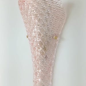 May include: A pink glass wall sconce with a textured, waffle-like pattern. The sconce has a cone shape and is mounted on a white wall.