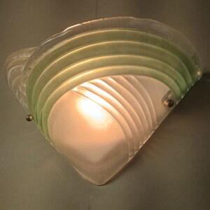 May include: A frosted glass wall sconce with a green glass overlay. The sconce has a geometric design with curved lines and a light bulb inside.