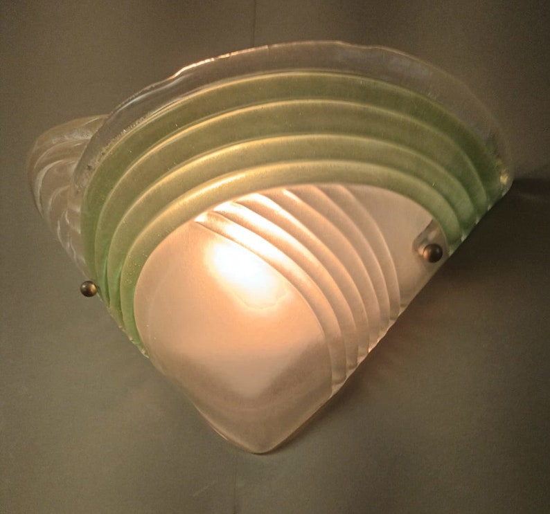 May include: A frosted glass wall sconce with a green glass overlay. The sconce has a geometric design with curved lines and a light bulb inside.