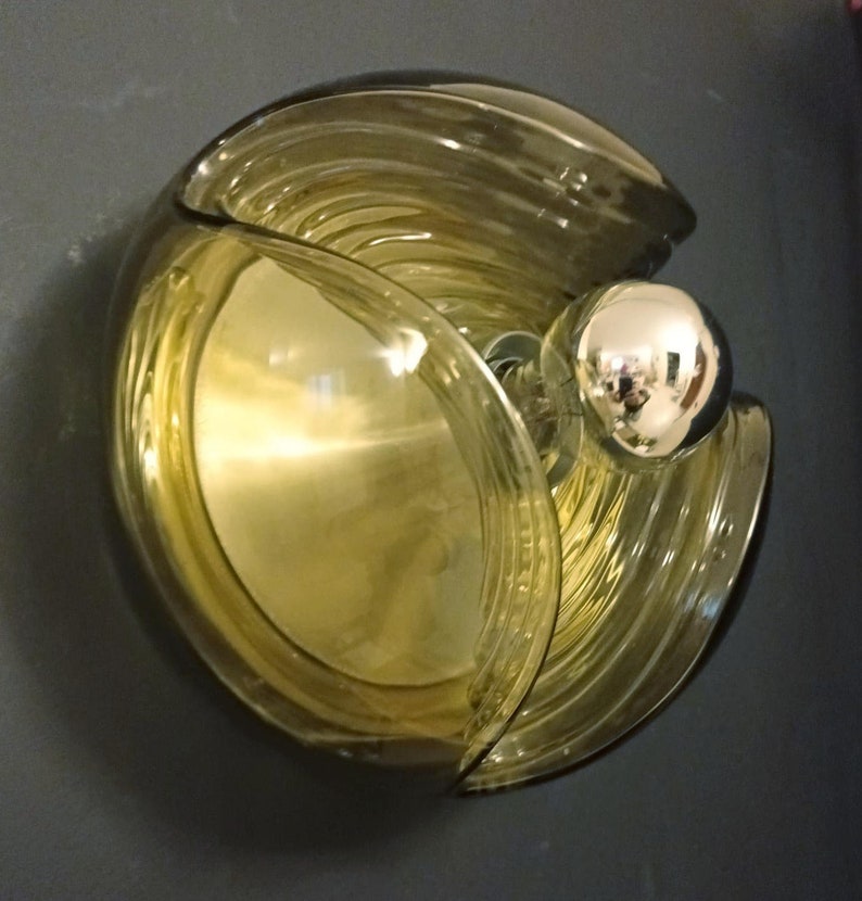 Puede incluir: Una l&aacute;mpara de pared de vidrio marr&oacute;n con un dise&ntilde;o redondo y en capas. La l&aacute;mpara tiene un accesorio de luz cromado y una bombilla de vidrio transparente.