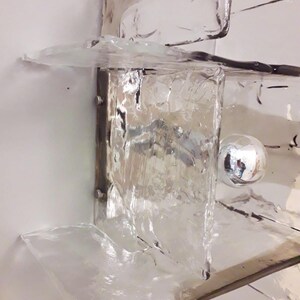 May include: A clear glass wall sconce with a textured surface and a silver metal base. The sconce has a round, silver light bulb in the centre.