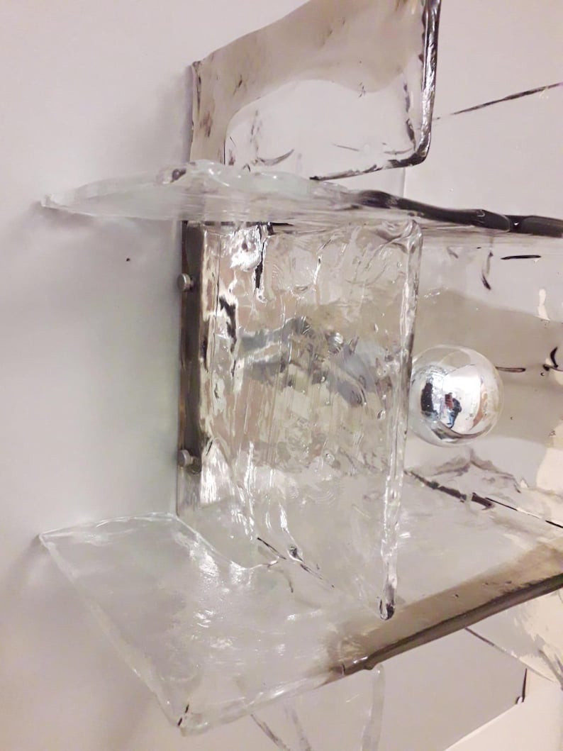 May include: A clear glass wall sconce with a textured surface and a silver metal base. The sconce has a round, silver light bulb in the centre.