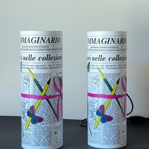 May include: Two white cylindrical lamps with a newspaper design. The lamps feature a pink ribbon, pencils, and a butterfly. The text on the lamps reads "IMMAGINARIO QUOTIDIANO ARTISTICO LETTERARIO SETTORE ARTISTICO, Via Baraldi, 14 Milano" and "ori nelle collezioni vedere in essi parentele con le sculture e le piccole cose - Nostalgia delle donne del Piemonte".