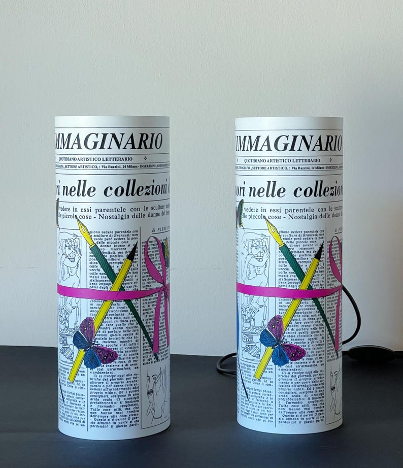 May include: Two white cylindrical lamps with a newspaper design. The lamps feature a pink ribbon, pencils, and a butterfly. The text on the lamps reads "IMMAGINARIO QUOTIDIANO ARTISTICO LETTERARIO SETTORE ARTISTICO, Via Baraldi, 14 Milano" and "ori nelle collezioni vedere in essi parentele con le sculture e le piccole cose - Nostalgia delle donne del Piemonte".