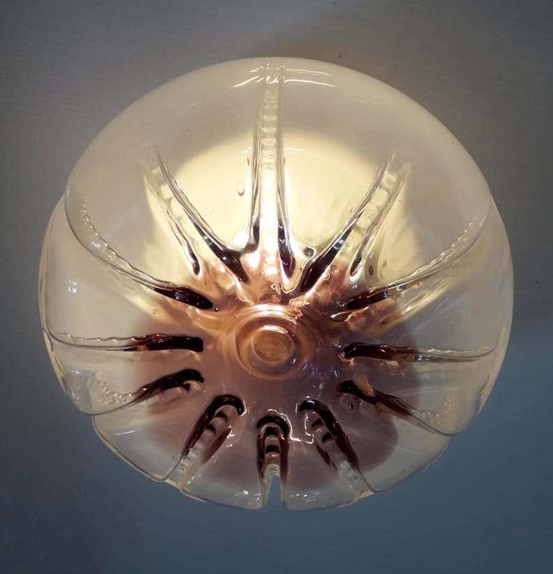 Pu&ograve; includere: Un paralume in plastica trasparente con un fondo marrone chiaro e testurizzato. Il paralume &egrave; illuminato dall'interno, creando una luce calda.