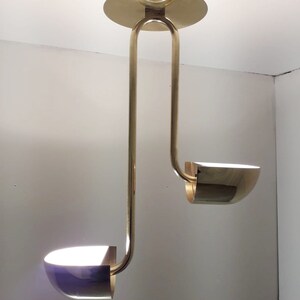 May include: A gold-toned, two-light pendant light fixture with a modern design. The fixture has two curved arms that extend downwards, each ending in a round, bowl-shaped light shade.