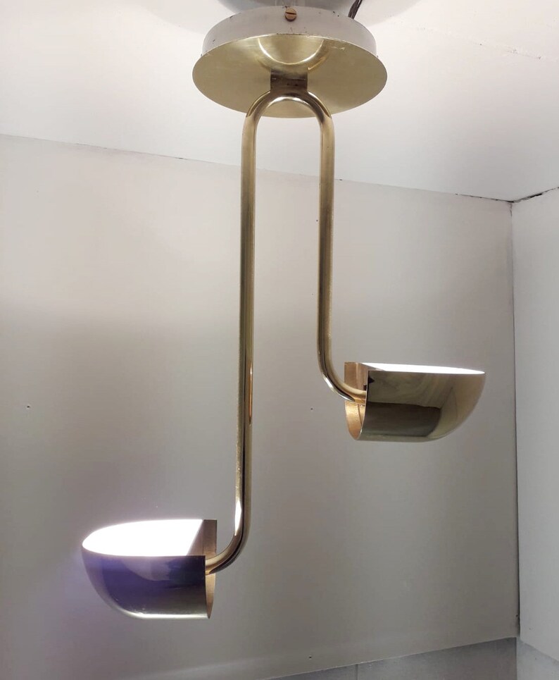 May include: A gold-toned, two-light pendant light fixture with a modern design. The fixture has two curved arms that extend downwards, each ending in a round, bowl-shaped light shade.
