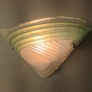 May include: A white and green glass wall sconce with a geometric design. The sconce is lit and casts a warm glow.