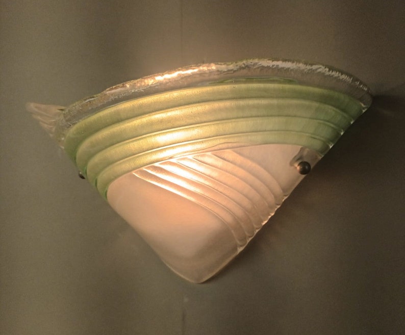 May include: A white and green glass wall sconce with a geometric design. The sconce is lit and casts a warm glow.