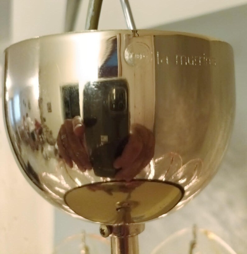 May include: A polished, metallic light fixture with a rounded, bowl-like shade. The surface reflects the surrounding environment, including a person holding a phone. The fixture has a decorative design and a central support rod.