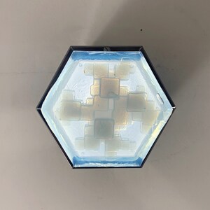 Może przedstawiać: Sześciokątna lampa sufitowa z czarną metalową ramą. Lampa posiada panel ze szkła mrożonego z wzorem kwadratowych kształt&oacute;w w odcieniach bieli i złota. Oprawa jest zamontowana na jasnoszarym suficie.