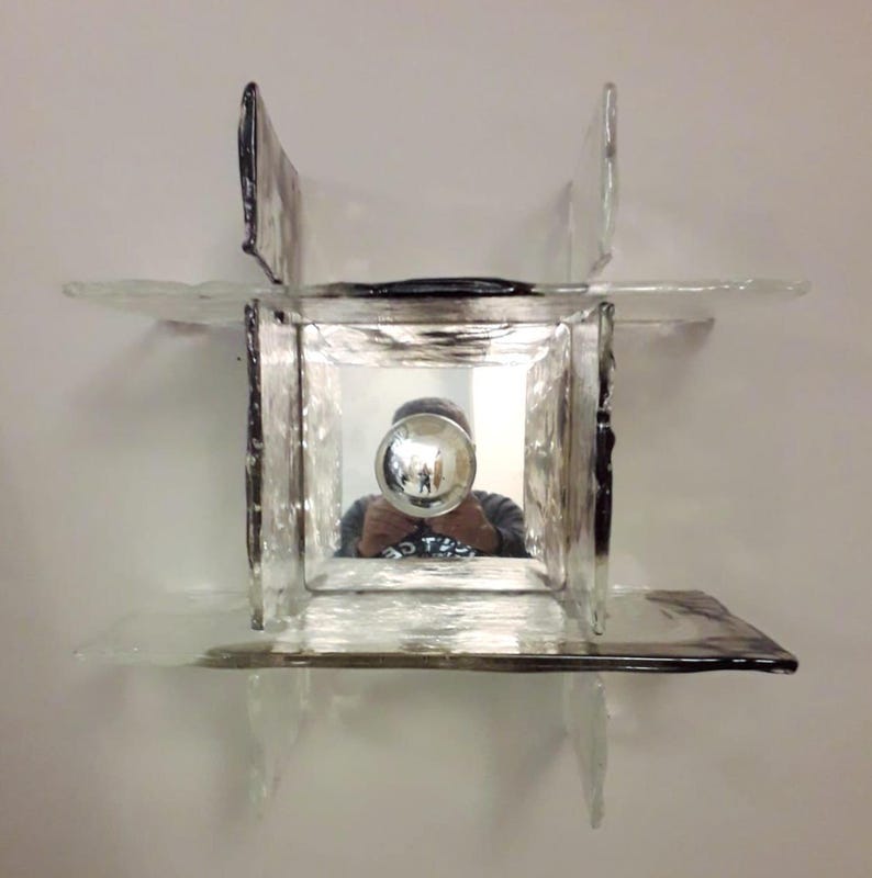 May include: A square-shaped glass wall sconce with a mirrored centre and a silver-coloured bulb. The sconce has a textured glass surface and is mounted on a white wall.