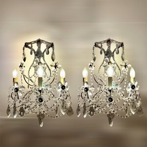 May include: Two ornate wall sconces with crystal accents. Each sconce features a dark metal frame with multiple light bulbs, crystal droplets, and floral embellishments. The sconces are designed to be mounted on a wall and provide ambient lighting.