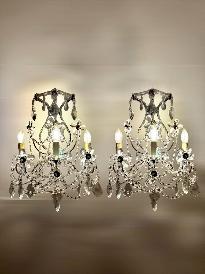 May include: Two ornate wall sconces with crystal accents. Each sconce features a dark metal frame with multiple light bulbs, crystal droplets, and floral embellishments. The sconces are designed to be mounted on a wall and provide ambient lighting.
