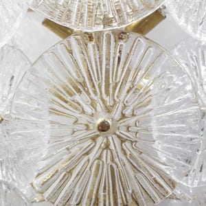 May include: Close-up of a vintage chandelier featuring clear, textured glass discs with gold-colored accents. The discs are arranged in a repeating pattern, connected by gold-toned metal rods and a central gold-colored stud.