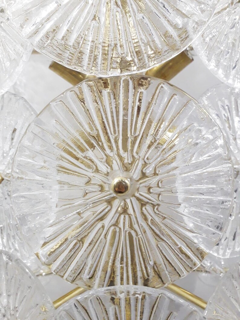 May include: Close-up of a vintage chandelier featuring clear, textured glass discs with gold-colored accents. The discs are arranged in a repeating pattern, connected by gold-toned metal rods and a central gold-colored stud.