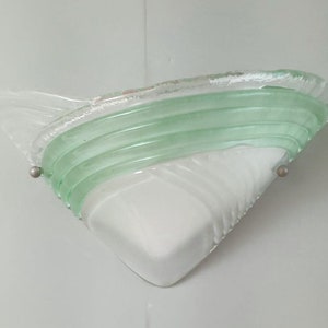 May include: A white and green glass wall sconce with a triangular shape and a wavy design. The sconce has a green glass band with a wavy design.