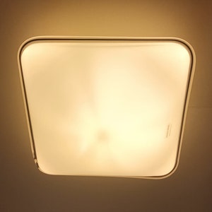 May include: A white square ceiling light fixture with a frosted glass cover. The light is turned on and emits a warm, yellow glow.