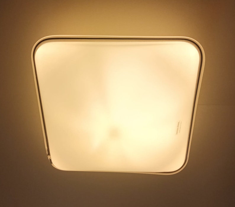 May include: A white square ceiling light fixture with a frosted glass cover. The light is turned on and emits a warm, yellow glow.