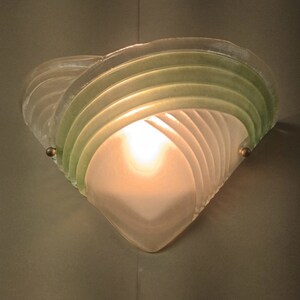 May include: A frosted glass wall sconce with a green glass overlay. The sconce has a geometric design and is lit by a single light bulb.