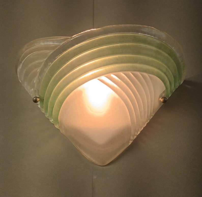 May include: A frosted glass wall sconce with a green glass overlay. The sconce has a geometric design and is lit by a single light bulb.