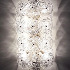 May include: A clear glass wall sconce with a series of circular, textured glass discs. Each disc has a black center and radiating lines, creating a starburst effect. The sconce is mounted on a metal frame, and the light shines through the glass.