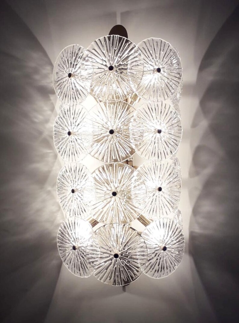 May include: A clear glass wall sconce with a series of circular, textured glass discs. Each disc has a black center and radiating lines, creating a starburst effect. The sconce is mounted on a metal frame, and the light shines through the glass.