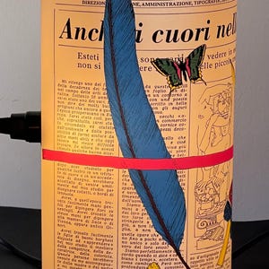 May include: A vintage newspaper clipping with a blue and black feather, a red and yellow butterfly, and the text "Anchi cuori nell'" in Italian. The newspaper is yellowed and has a vintage aesthetic.