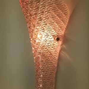 May include: A pink glass wall sconce with a textured, rippled design. The sconce is lit and casts a warm glow.