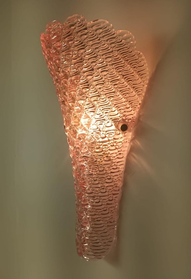May include: A pink glass wall sconce with a textured, rippled design. The sconce is lit and casts a warm glow.