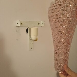 May include: A pink glass wall sconce with a textured bubble design. The sconce is attached to a white metal bracket with a light bulb socket.