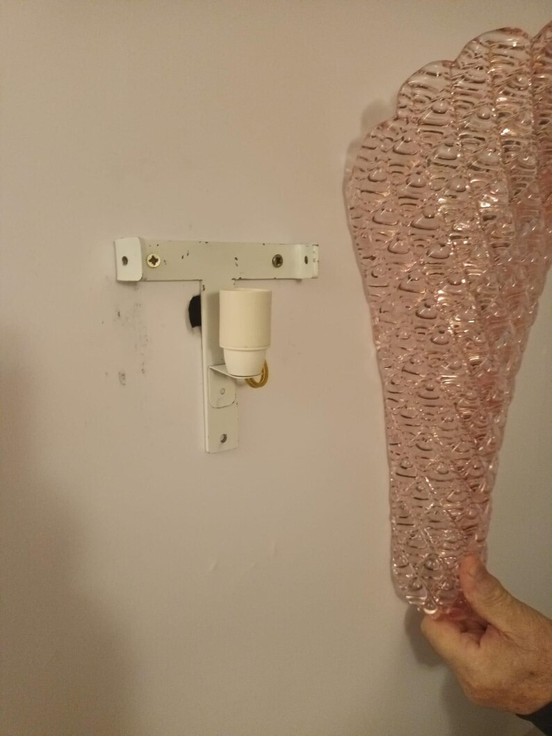 May include: A pink glass wall sconce with a textured bubble design. The sconce is attached to a white metal bracket with a light bulb socket.