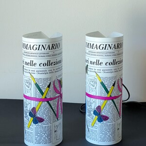 May include: Two white cylindrical lamps with a newspaper design. The lamps feature a pink ribbon, pencils, and a butterfly graphic. The text on the lamps reads "Imaginario Quotidiano Artistico Letterario".