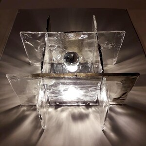 May include: A modern wall sconce with a geometric design. The sconce is made of clear glass panels with a textured surface. A light bulb is visible inside the sconce.