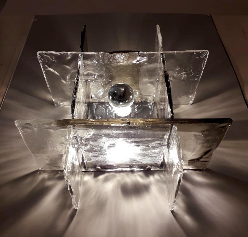 May include: A modern wall sconce with a geometric design. The sconce is made of clear glass panels with a textured surface. A light bulb is visible inside the sconce.