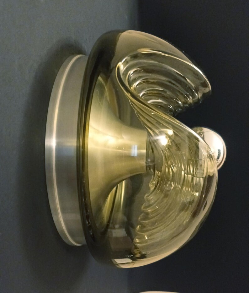 Puede incluir: Una l&aacute;mpara de pared de vidrio marr&oacute;n con una base de metal plateado. El vidrio tiene una forma ondulada, org&aacute;nica y una superficie texturizada.