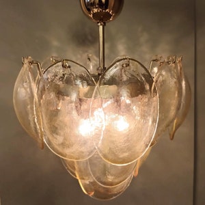 May include: A vintage chandelier with a gold-toned metal frame and multiple petal-shaped glass shades. The glass shades are a light amber color and have a textured surface. The light fixture is suspended from a gold-colored ceiling mount.
