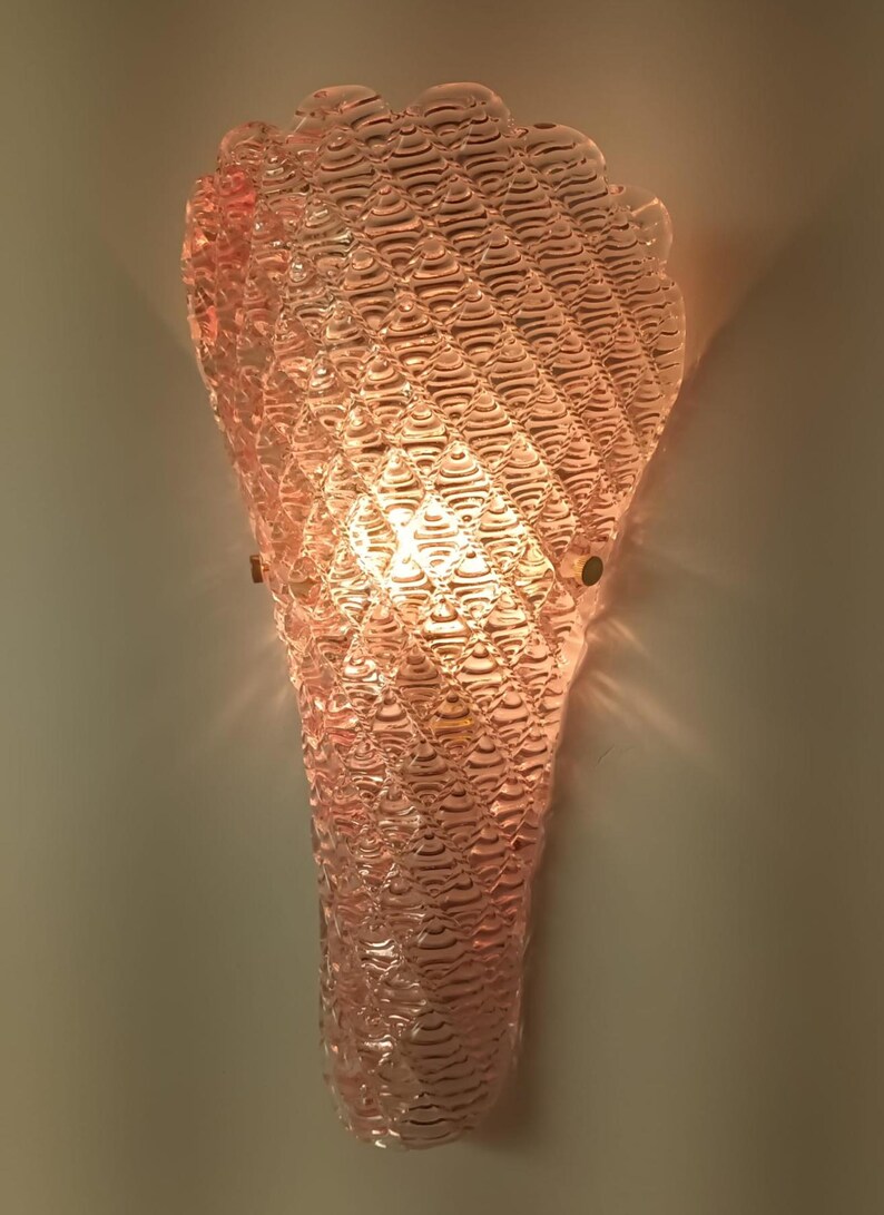 May include: A pink glass wall sconce with a textured, bubble-like design. The sconce is lit with a light bulb, casting a warm glow.