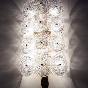 May include: A decorative wall sconce featuring multiple clear, circular glass elements arranged in a layered design. Each disc has a textured, radial pattern and a dark center. The sconce has a gold-toned metal frame and a dark, oval base.
