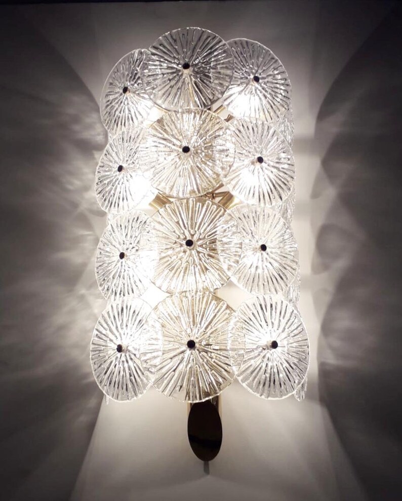 May include: A decorative wall sconce featuring multiple clear, circular glass elements arranged in a layered design. Each disc has a textured, radial pattern and a dark center. The sconce has a gold-toned metal frame and a dark, oval base.