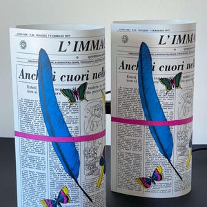 May include: Two decorative lamps shaped like rolled-up newspapers with a blue feather, butterflies, and pink and yellow accents. The text on the newspaper reads "L'IMMAGINE" and "Anchi cuori nelle".