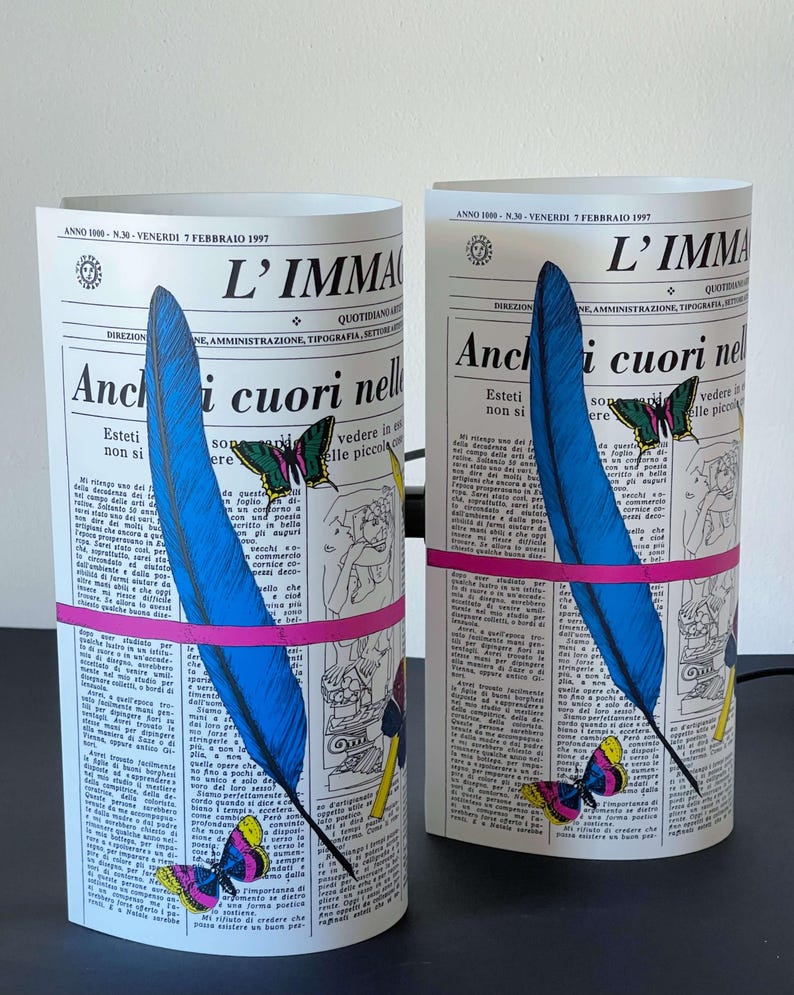 May include: Two decorative lamps shaped like rolled-up newspapers with a blue feather, butterflies, and pink and yellow accents. The text on the newspaper reads "L'IMMAGINE" and "Anchi cuori nelle".