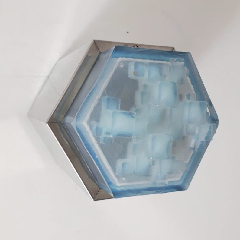 Może przedstawiać: Sześciokątny element artystyczny ze srebrną metalową ramą. Wnętrze ma jasnoniebieski, geometryczny wz&oacute;r o teksturowanym wyglądzie. Element jest zamontowany na białej ścianie.