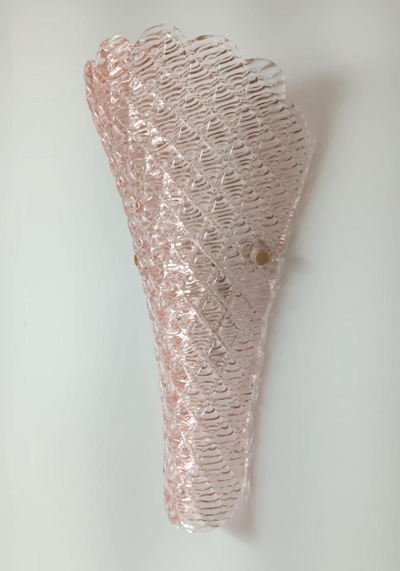 May include: A pink glass wall sconce with a textured, wavy design. The sconce has a cone shape and is attached to the wall with two gold-colored brackets.
