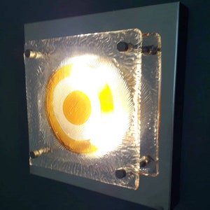 May include: A square wall sconce with a frosted glass shade featuring a concentric circle design in shades of yellow and white. The light is on and illuminating the room.