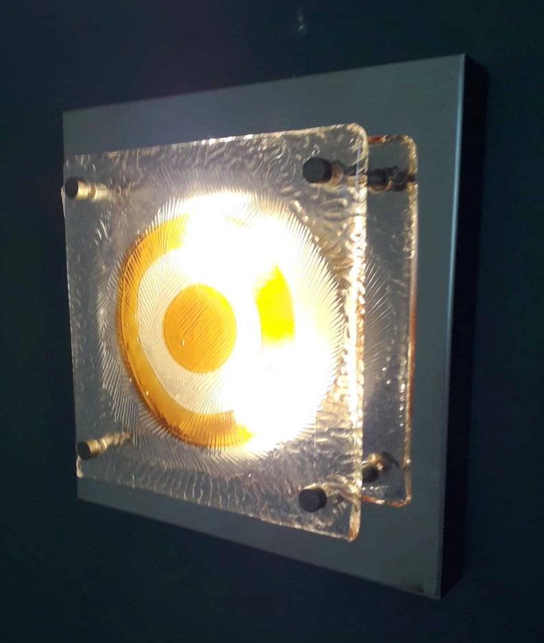 May include: A square wall sconce with a frosted glass shade featuring a concentric circle design in shades of yellow and white. The light is on and illuminating the room.