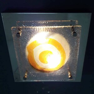 May include: A square ceiling light fixture with a frosted glass shade featuring a concentric circle design in shades of yellow and white.