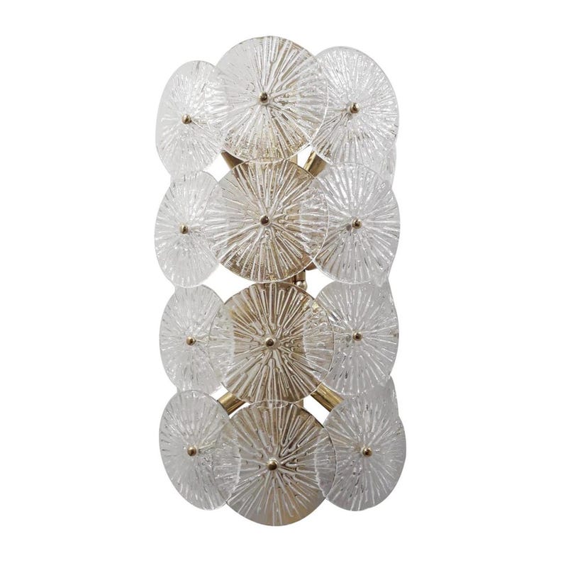 May include: A decorative wall sconce featuring a series of clear, circular glass elements with radial patterns. The glass circles are arranged in a vertical column and are connected by a gold-colored metal frame. Each glass element has a small, gold-colored center.