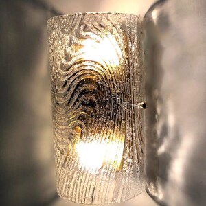 May include: A gold-toned wall sconce with a clear glass shade featuring a textured, wavy pattern. The light is on and illuminating the room.