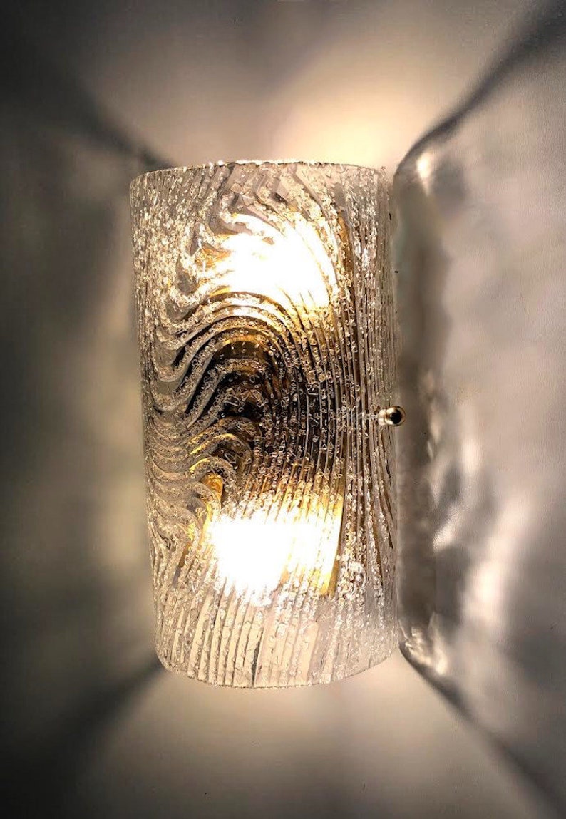 May include: A gold-toned wall sconce with a clear glass shade featuring a textured, wavy pattern. The light is on and illuminating the room.